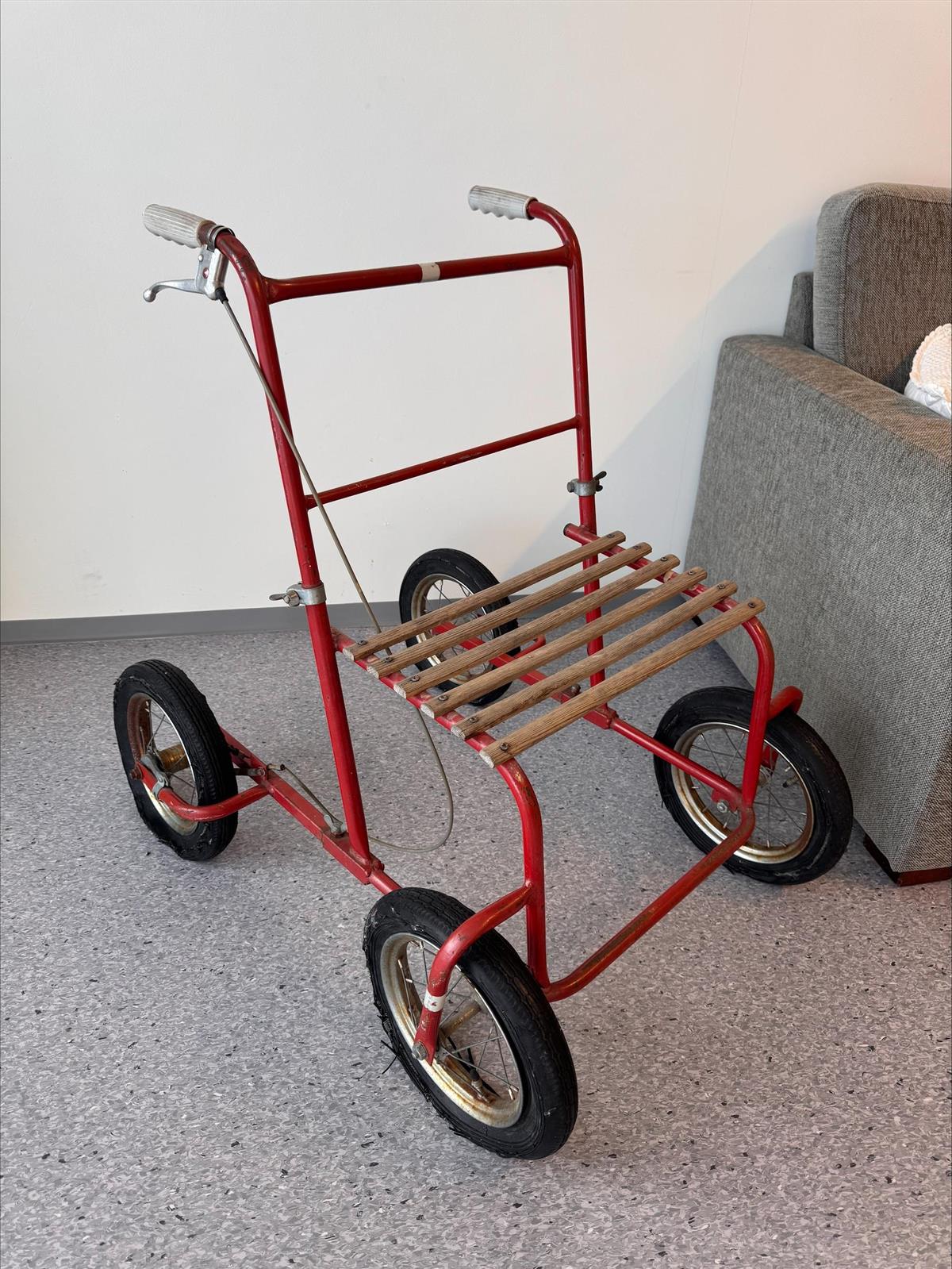 Rød rullator på et grått gulvbelegg foran en lys vegg - Klikk for stort bilde